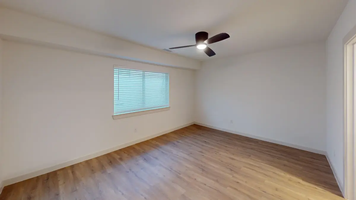 Empty bedroom with warm cream walls, a bronze ceiling fan, a blinded window on the left, and honey-toned wood-look flooring