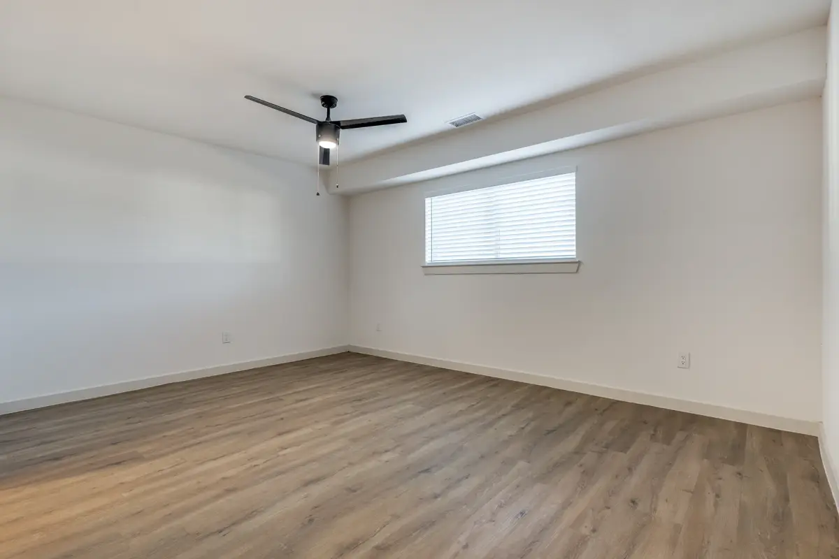 Empty bedroom with bright white walls, a black ceiling fan, a ceiling vent, a blinded window on the right, and gray-toned wood-look flooring