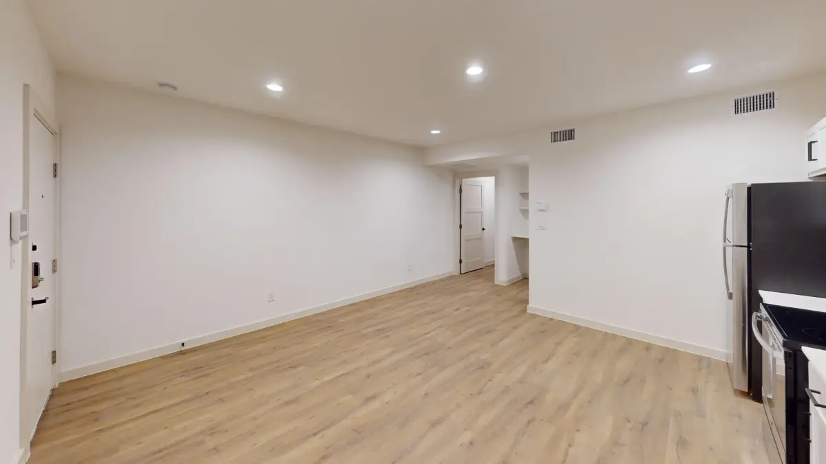 Empty open living area with wood-look flooring opening to a hallway and the kitchen