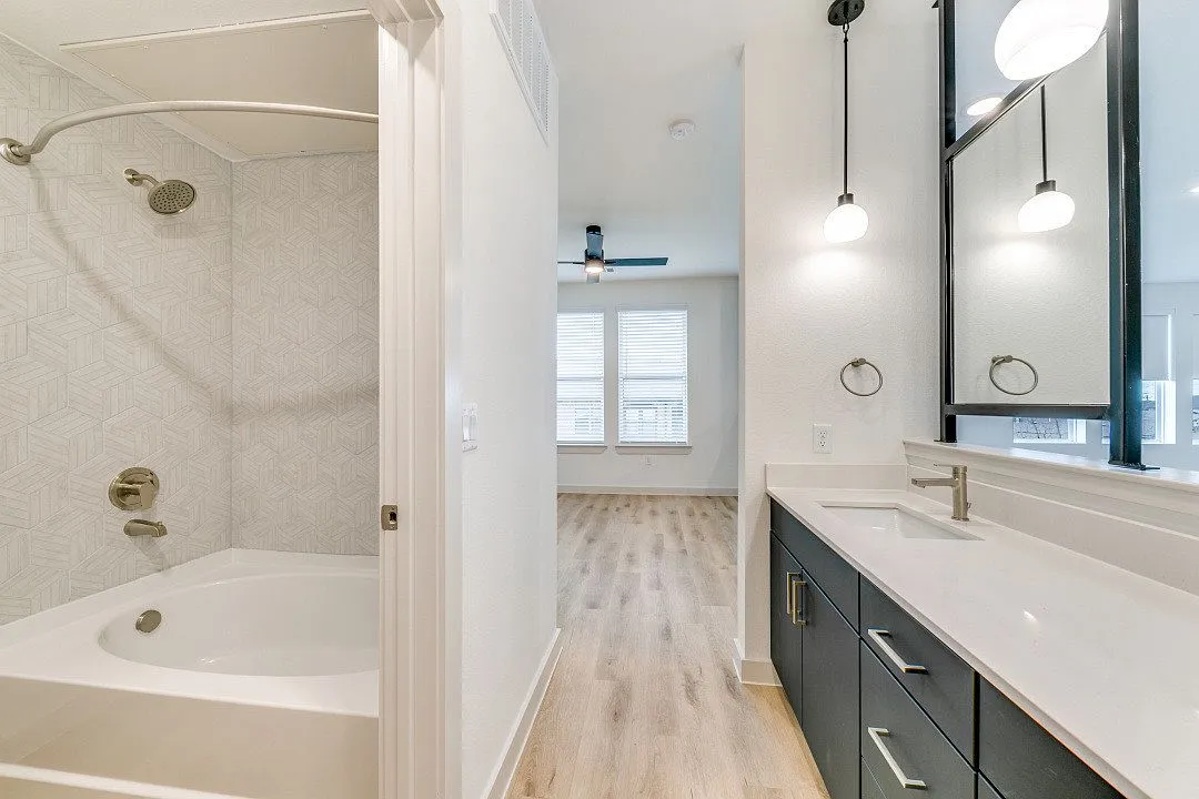 En-suite bath with a garden tub set in geometric mosaic tile beside a long white-quartz vanity with dark cabinetry and globe pendant lights, opening to the bedroom