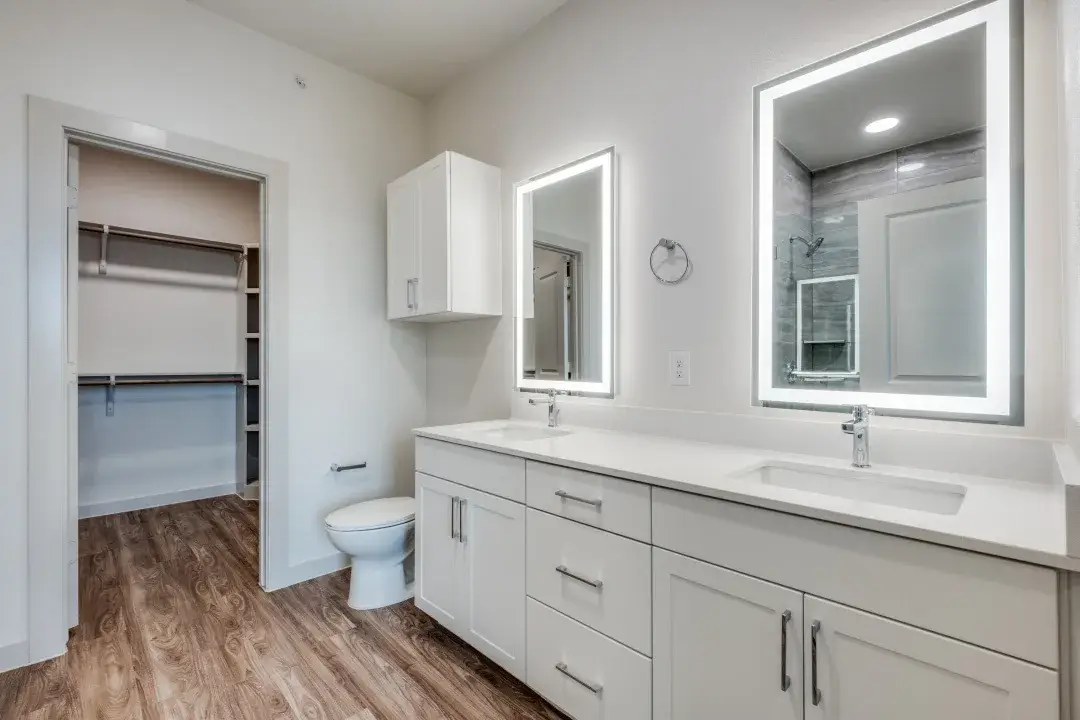 Bathroom with a white double-sink vanity, white quartz top, two backlit mirrors, and an adjoining walk-in closet