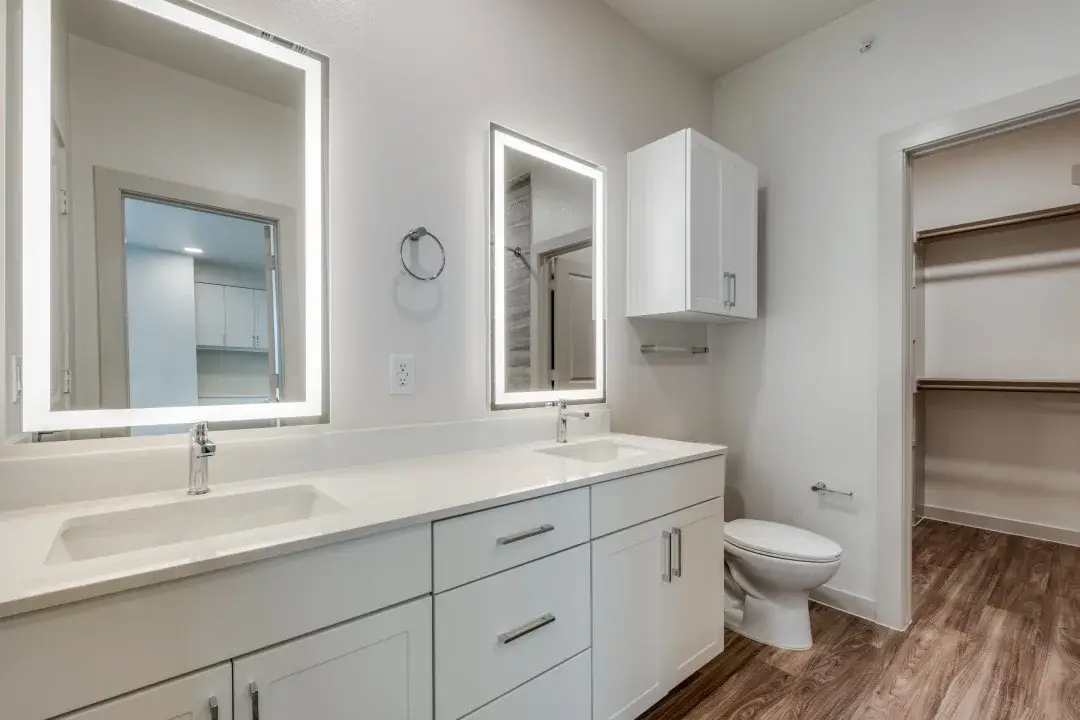 Bathroom with a white double vanity, two backlit LED mirrors, an upper cabinet, and a walk-in closet beyond