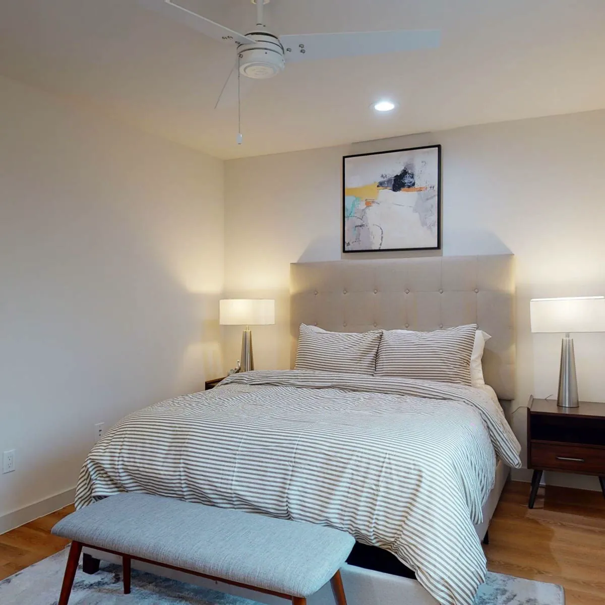 Bedroom with tufted beige headboard, striped duvet, matching bedside lamps, ceiling fan, and an upholstered bench