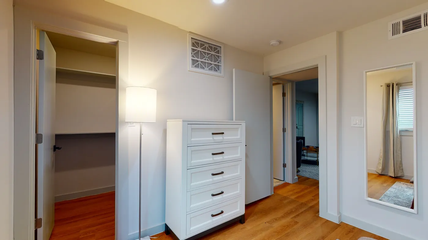Bedroom corner with a white five-drawer dresser, drum floor lamp, open walk-in closet, and full-length wall mirror