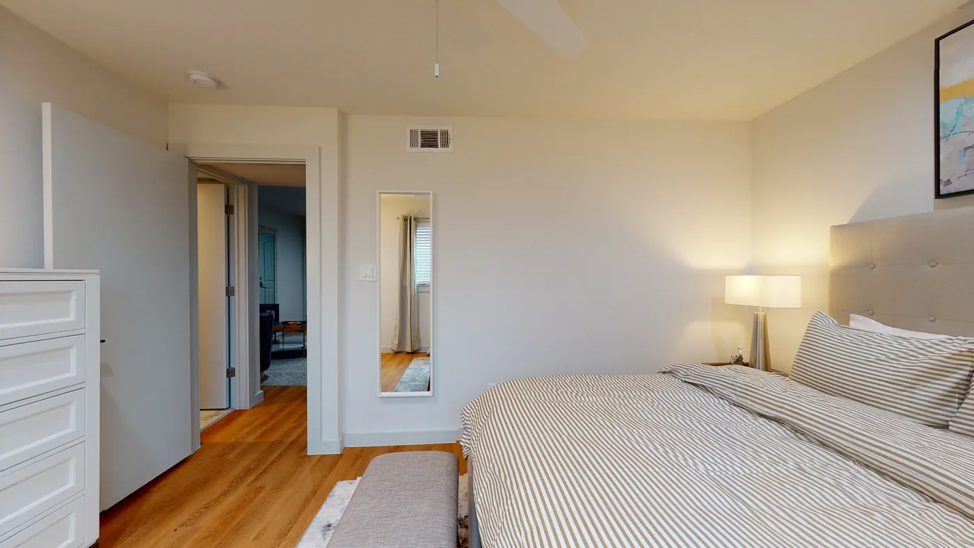 Striped duvet and tufted headboard beside a slim wall mirror, with wood-look floors leading toward the living area