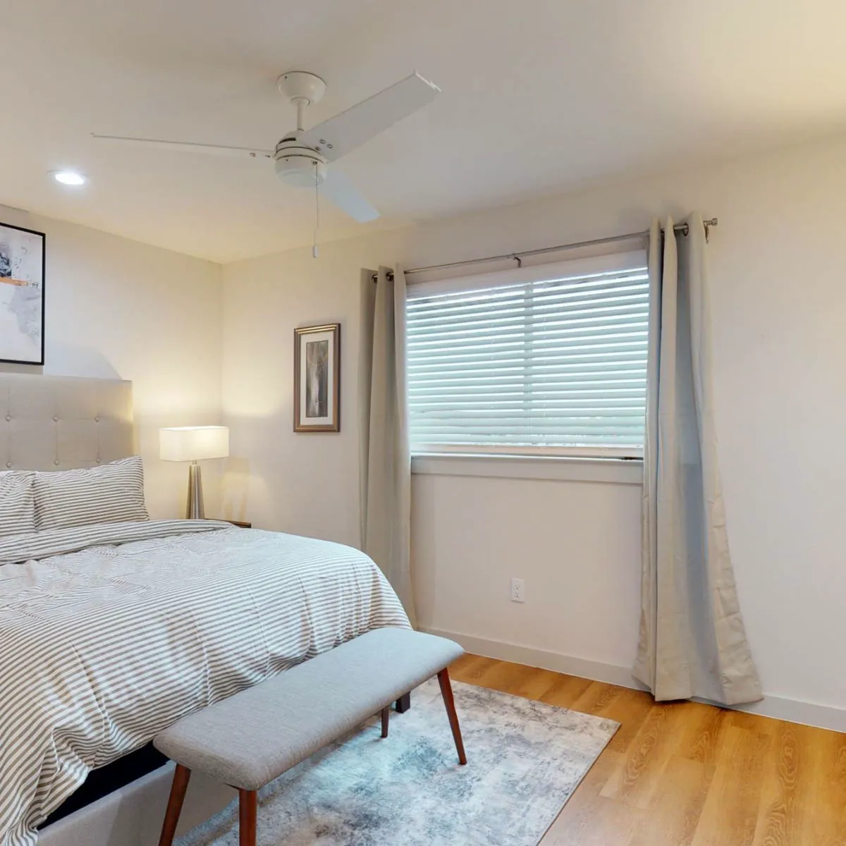 Bright bedroom with a ceiling fan, wide blinds framed by floor-length curtains, and a bench at the foot of the bed
