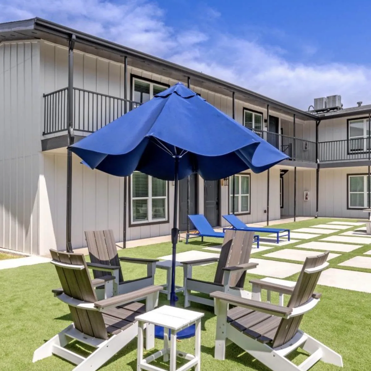 Navy umbrella over gray Adirondack chairs on turf, with blue loungers along the balcony-lined two-story courtyard
