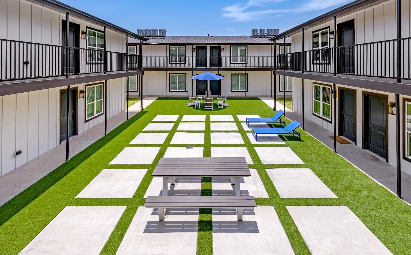 Checkerboard pavers and turf between balcony walkways, with a picnic table, blue loungers, and umbrella seating