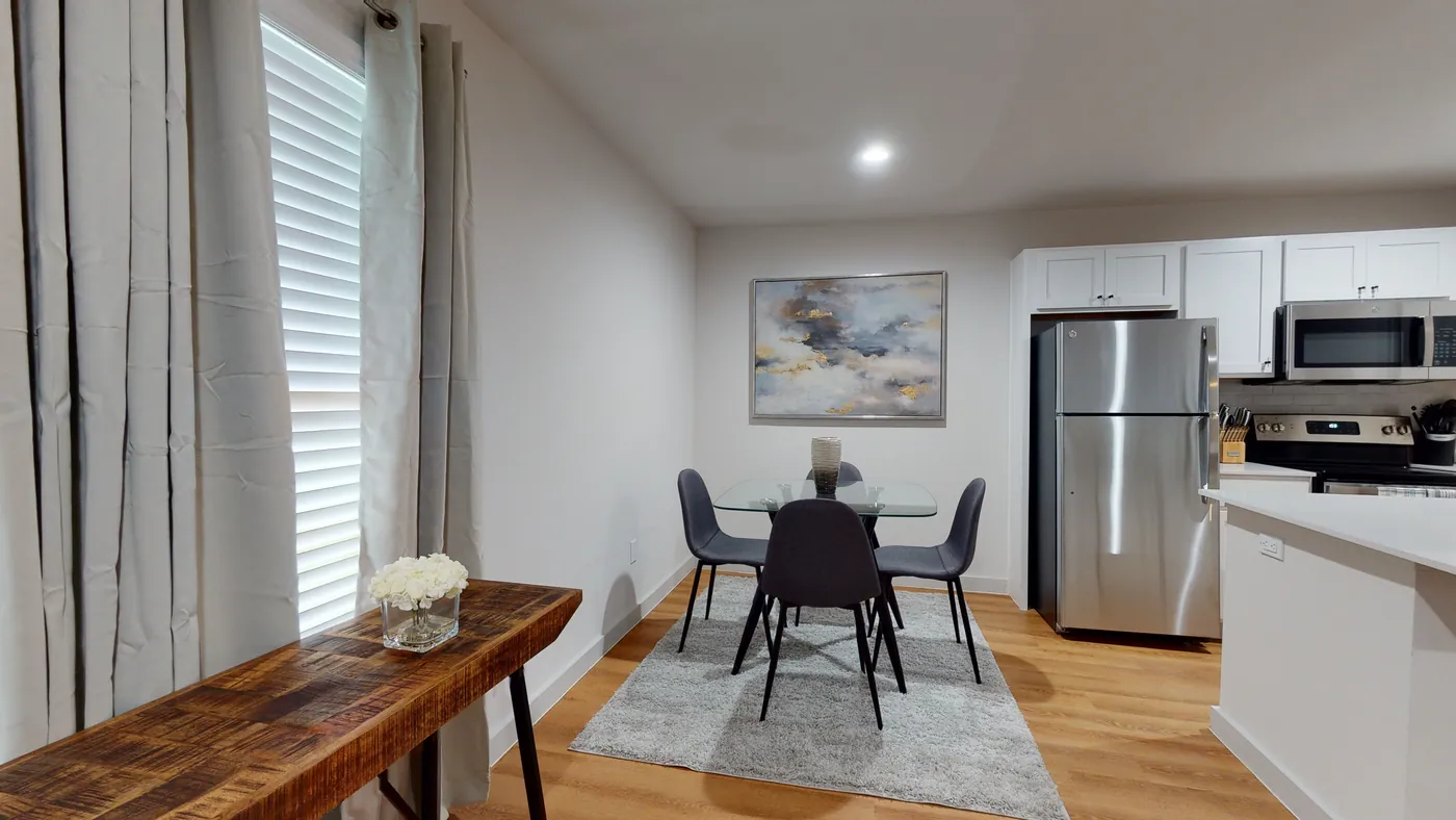 Glass-top dining table with four gray chairs on a plush rug, beside a rustic wood console and stainless refrigerator