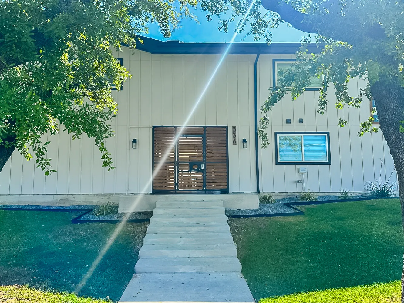 White board-and-batten facade of Hoskins 90 with a wood-slat entry gate, black lanterns, and oak-shaded front lawn