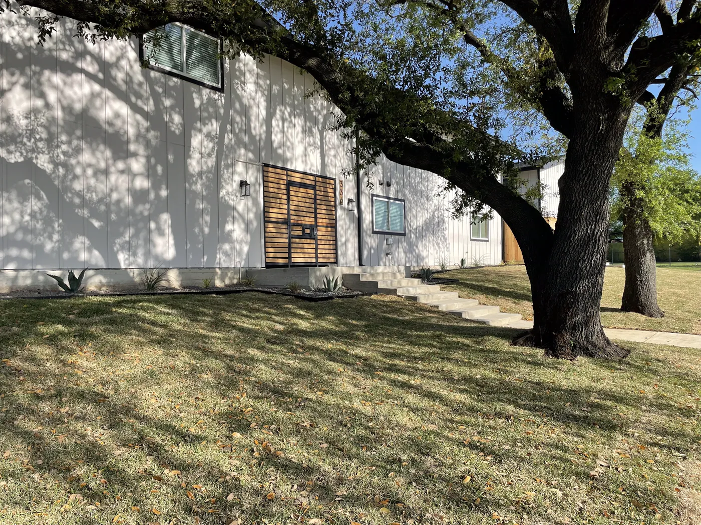 Slatted-wood entry gate on white vertical siding, shaded by a mature oak with agave plantings along the foundation