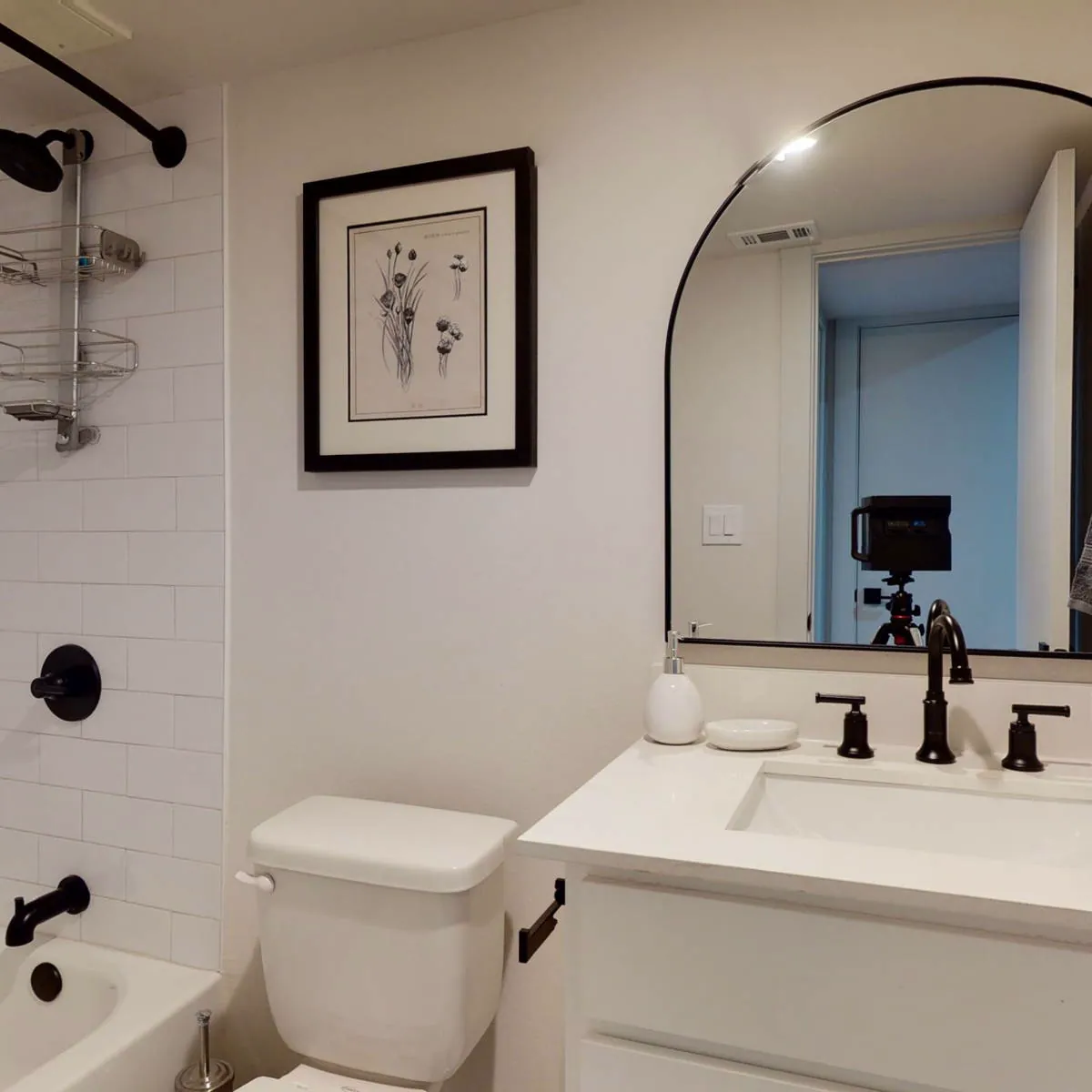 Bathroom with white subway-tile tub surround, matte-black fixtures, arched black-framed mirror, and quartz vanity