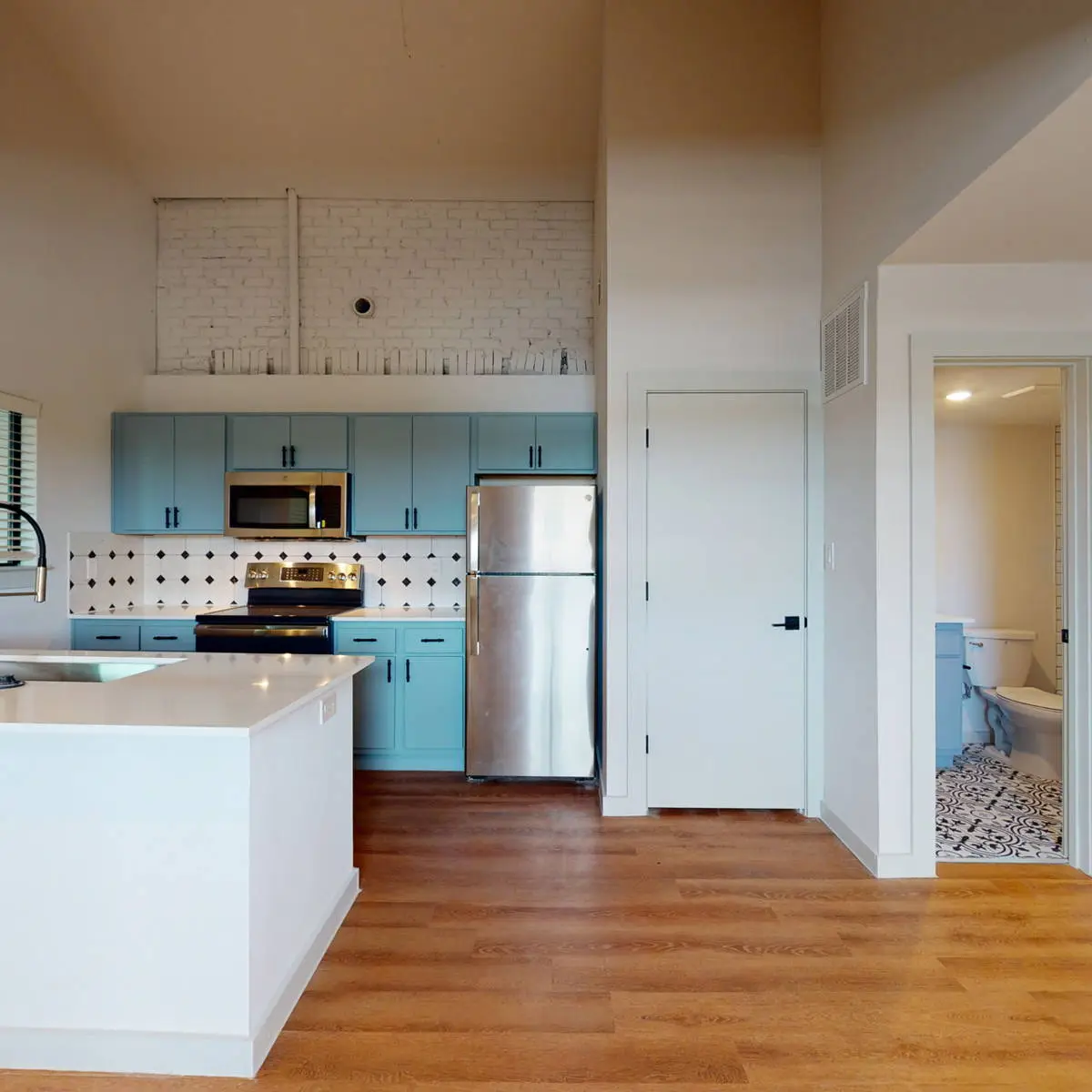 Loft kitchen with teal cabinets and a quartz peninsula under a vaulted ceiling, opening to a bathroom with patterned floor tile