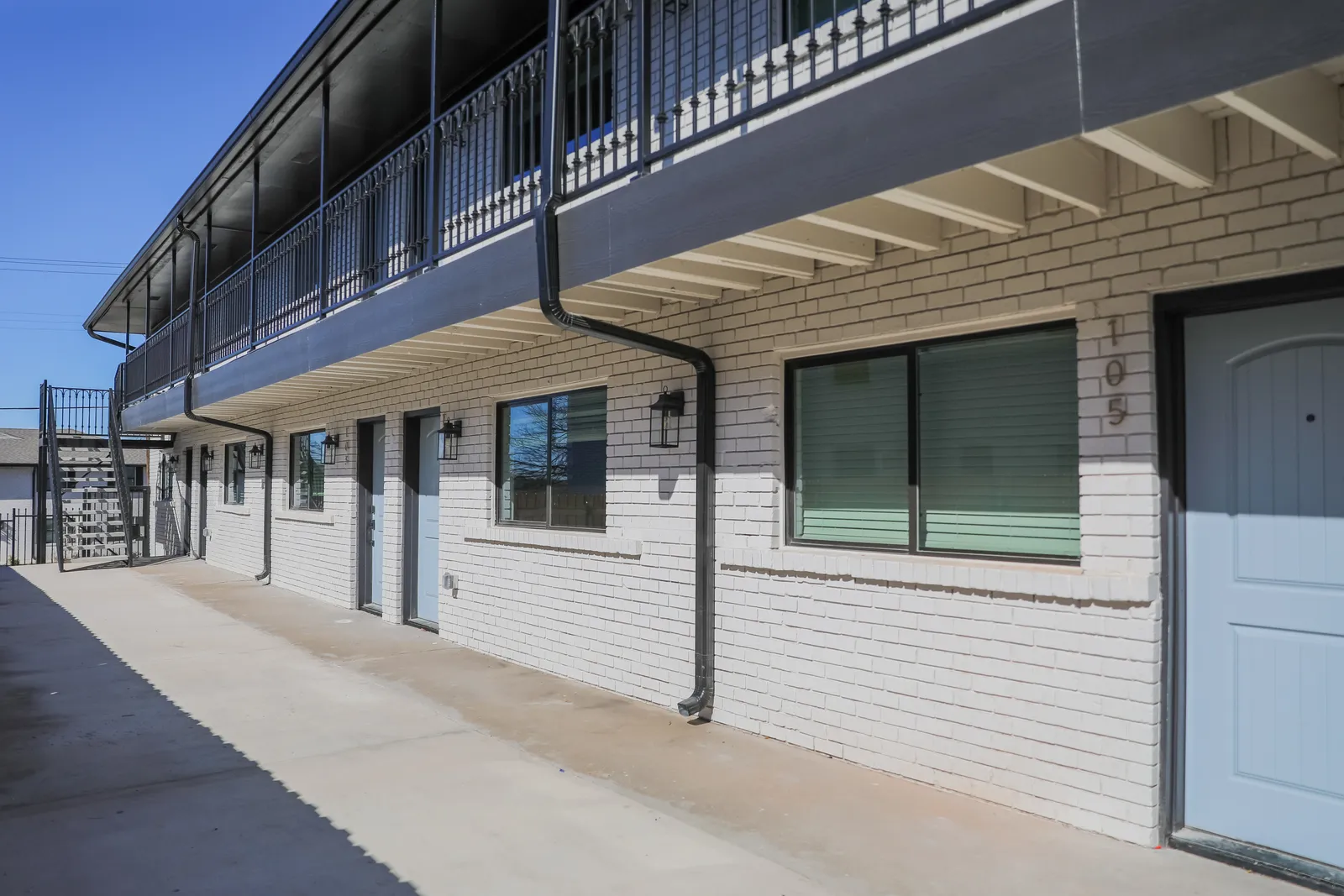 White-painted brick building with a black steel balcony walkway, lantern-lit ground-floor entries, and stair access