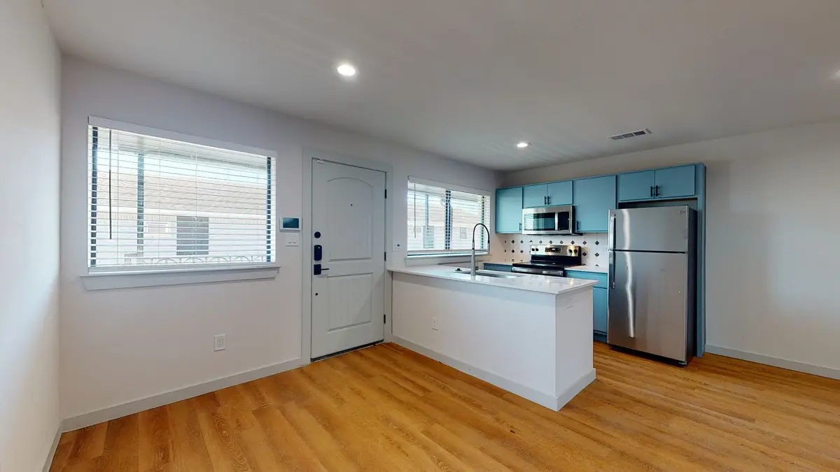Open kitchen with teal cabinets and stainless appliances and a quartz peninsula beside a light-blue entry door