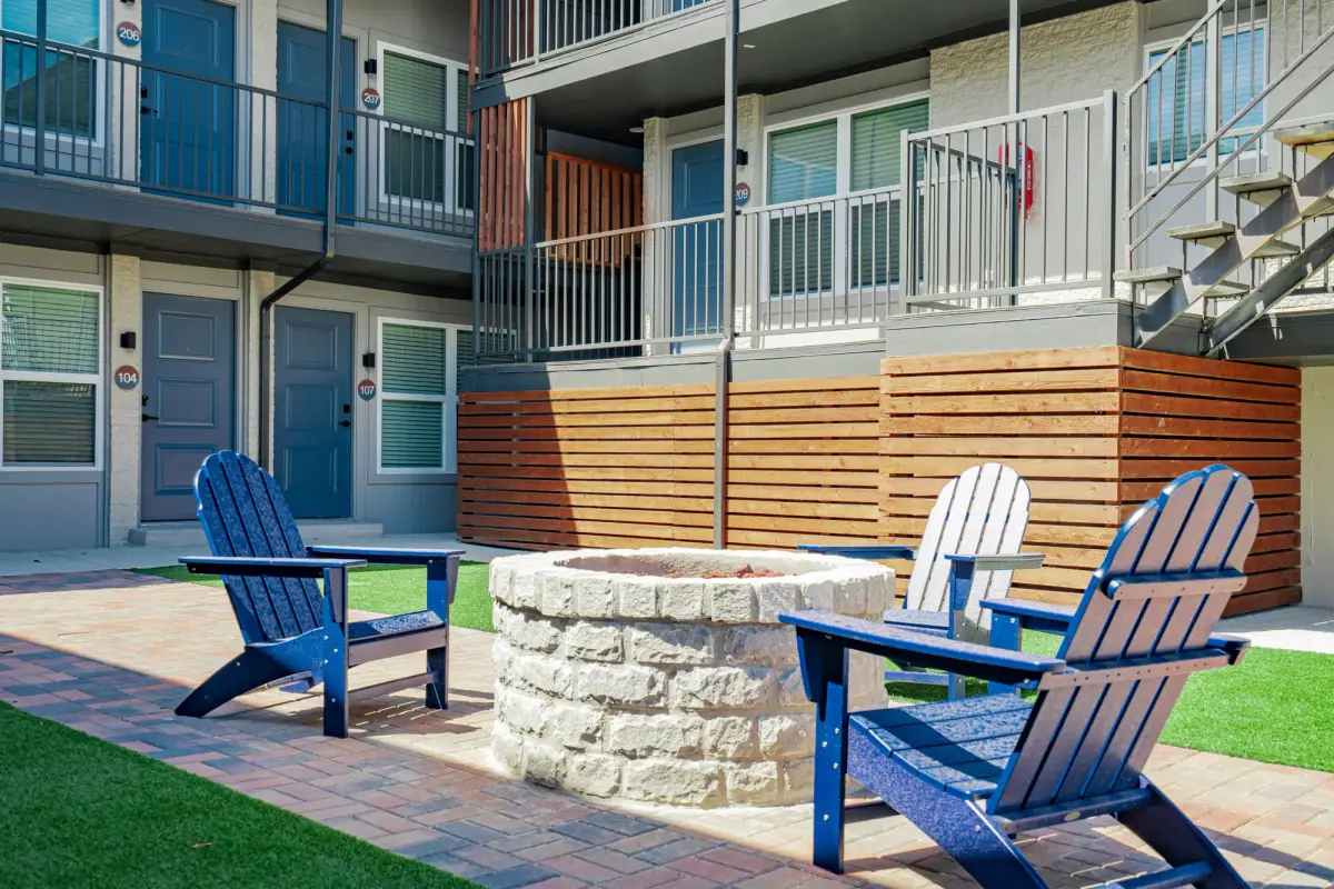 Navy Adirondack chairs circle a round stone fire pit against cedar-slat skirting below ground-floor unit doors