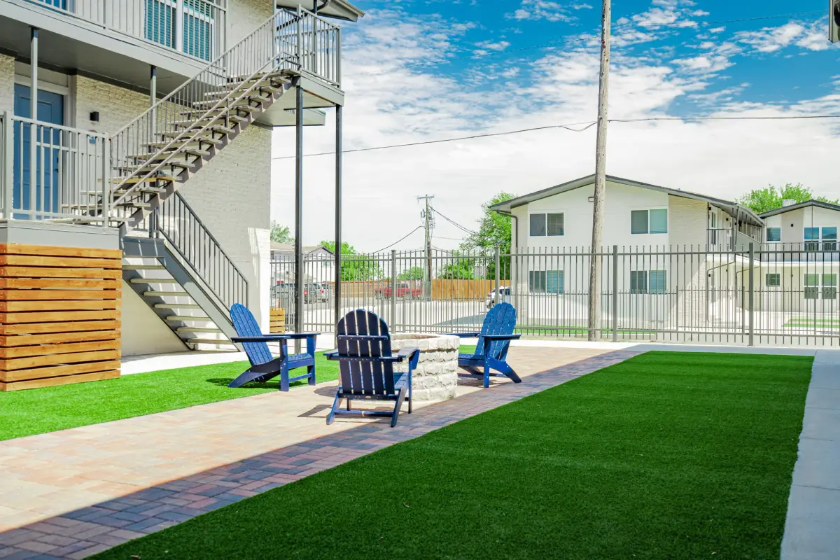 Blue Adirondack chairs and a stone fire pit on turf and pavers, with an exterior stair and iron perimeter fence