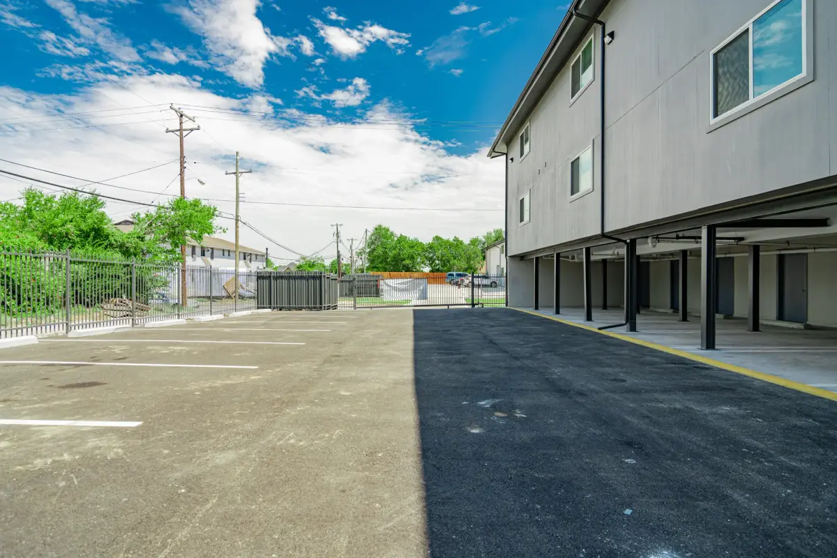 Striped open parking lot beside covered spaces under the building, ringed by a black iron fence and sliding gate