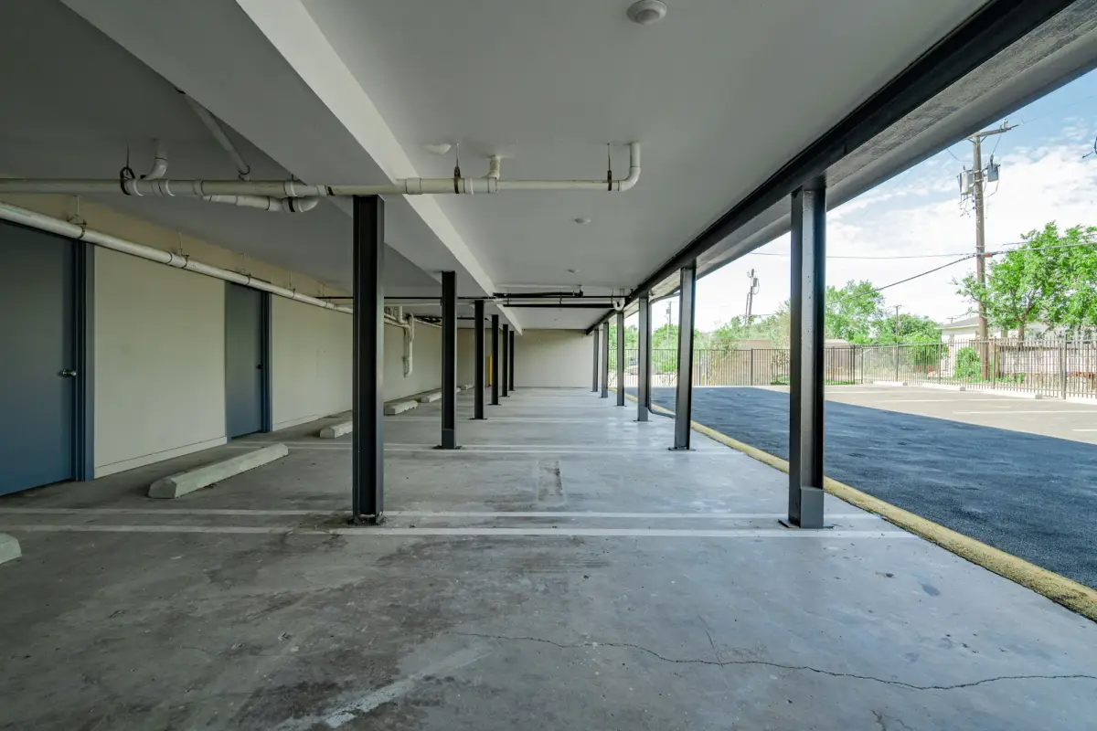 Covered parking beneath the building with steel columns, concrete wheel stops, striped bays, and gray storage doors