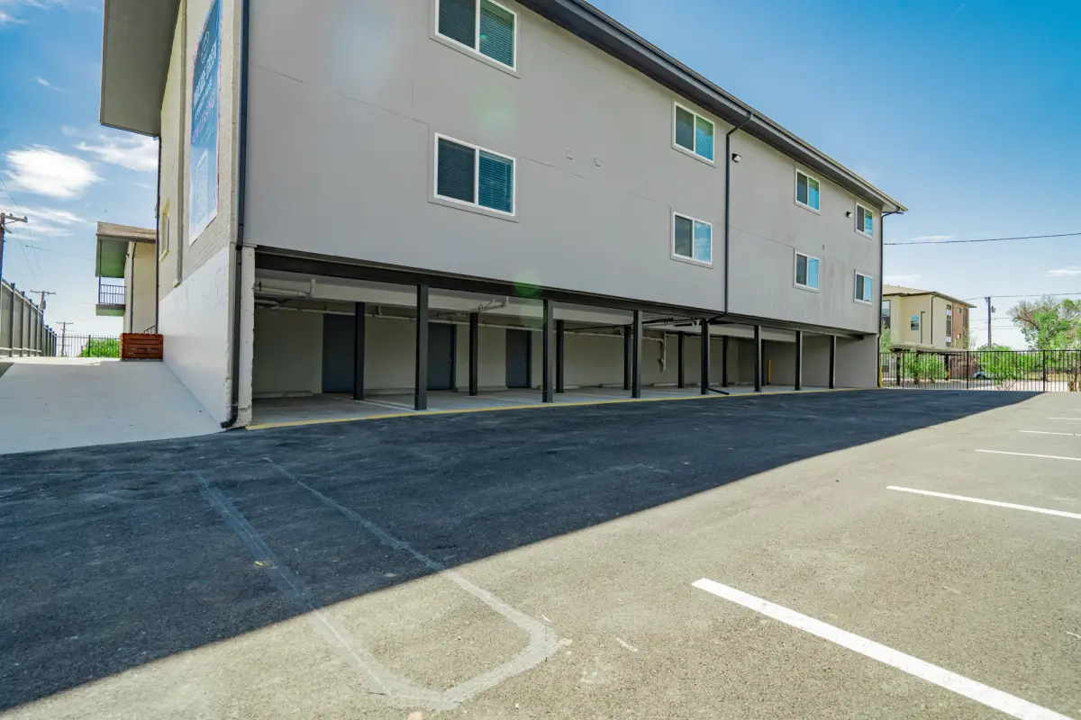 Tuck-under covered parking bays along a gray building, with freshly paved asphalt drive and white-striped spaces