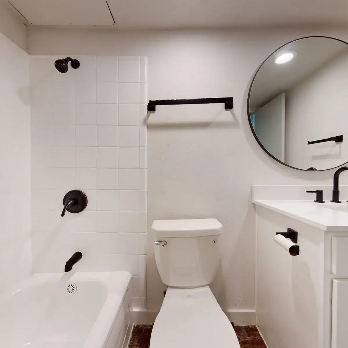 Bathroom with a white vanity, granite top, black fixtures, a round black-framed mirror, and a white-tile tub-and-shower