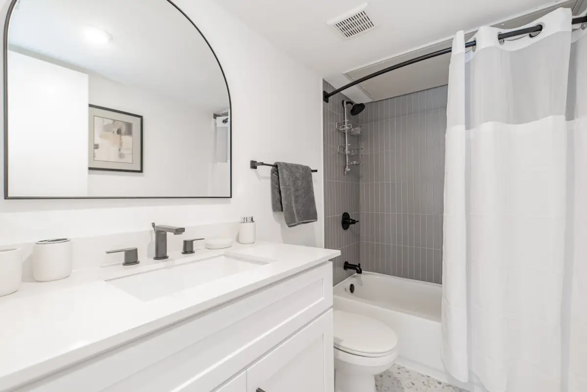 Bathroom with a white vanity, white quartz top, black fixtures, an arched mirror, and a gray-tile tub-and-shower