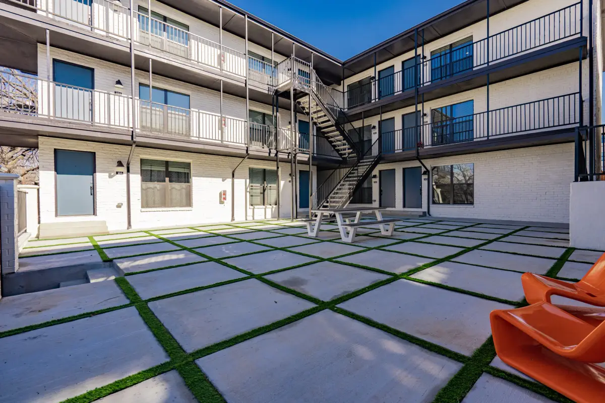 Interior courtyard with a paver-and-turf grid, a picnic table, and orange molded lounge chairs below three-story balcony walkways