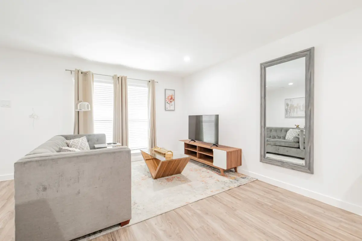 Living room with a gray sofa, media console and TV, a leaning mirror, and a curtained window