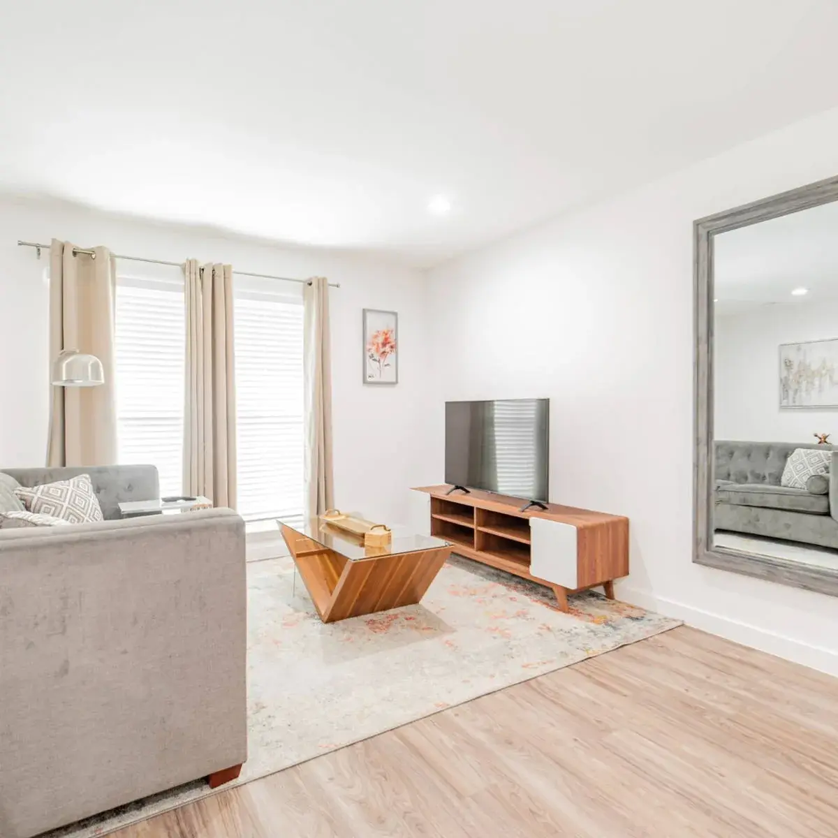 Living room with a gray sofa, wood media console and TV, a leaning mirror, and a curtained window