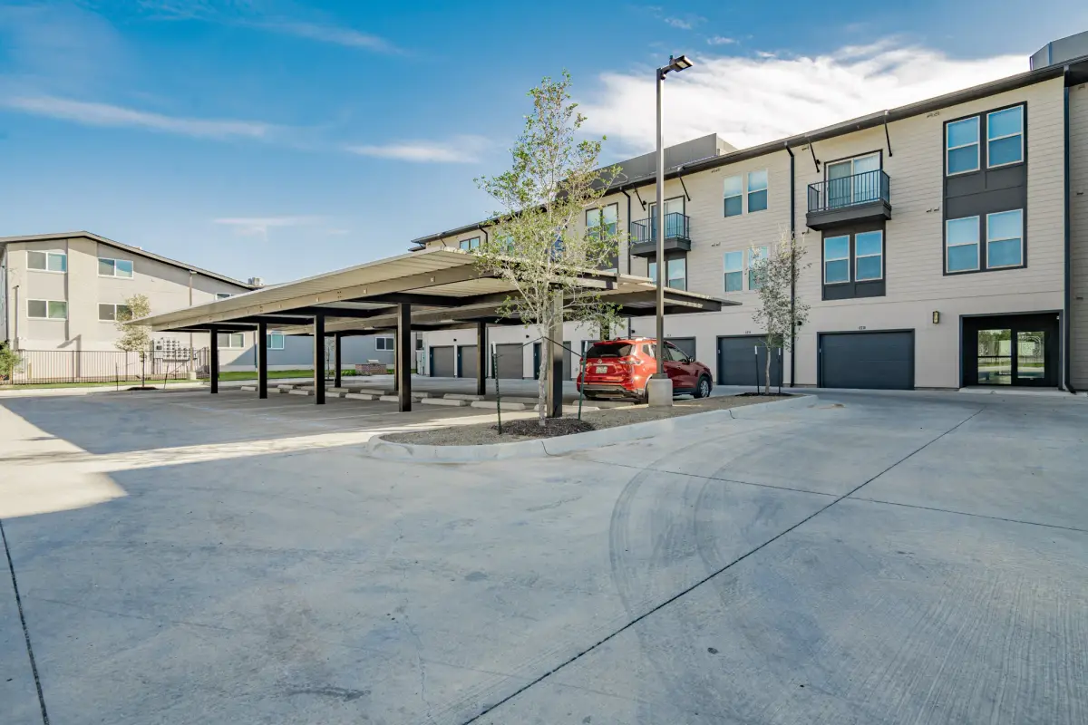 Steel-frame carport canopy in the open concrete lot, with garage doors and balconies on the building behind