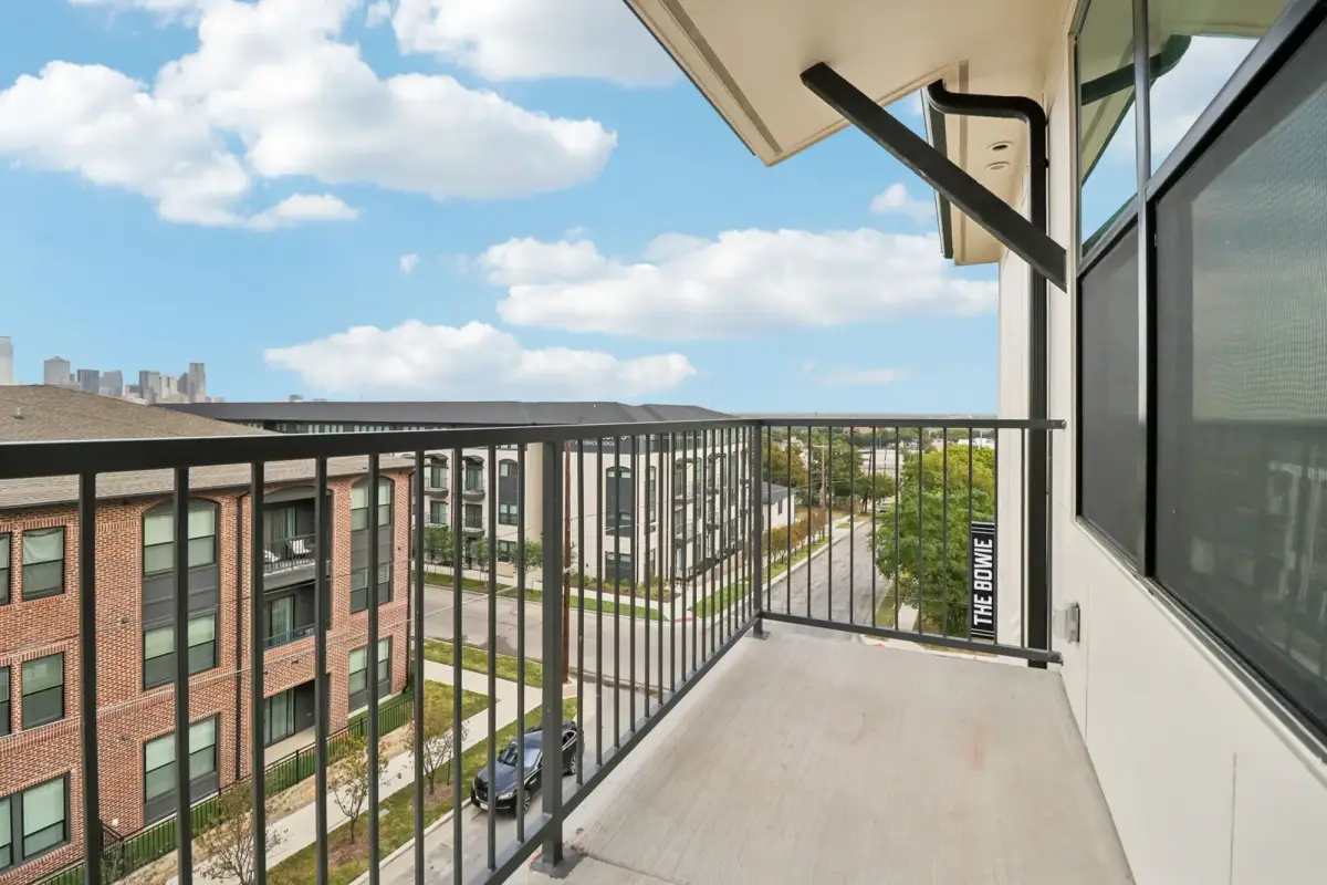Balcony with black metal railing overlooking a tree-lined street, The Bowie blade sign, and the Dallas skyline