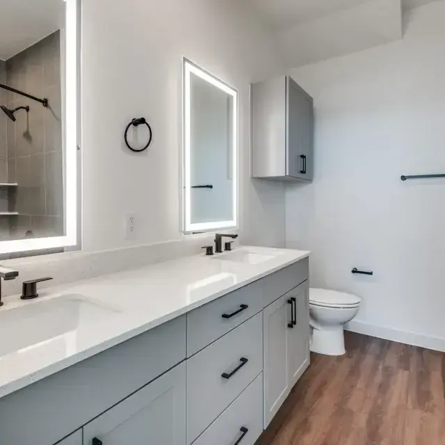 Bathroom with a gray double-sink vanity, white quartz top, backlit mirrors, black fixtures, and a tiled tub-and-shower