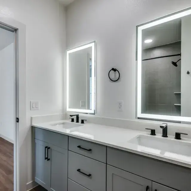 Bathroom with a gray double vanity, two backlit LED mirrors, black fixtures, and a tiled walk-in shower
