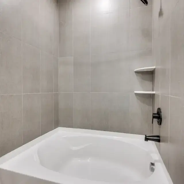 Tub-and-shower with a gray tile surround, white tub, corner shelves, and a black fixture set