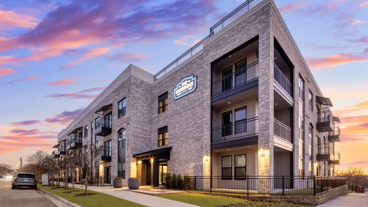 The Cambridge at dusk, three stories of light brick with black metal balconies, a lit nameplate on the corner, and a landscaped walkway to the entrance