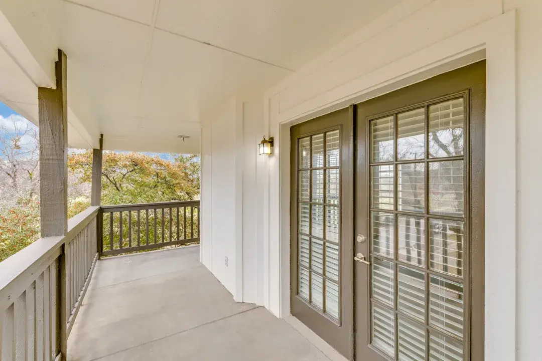 Private covered balcony with a wood railing and French doors with plantation shutters overlooking trees