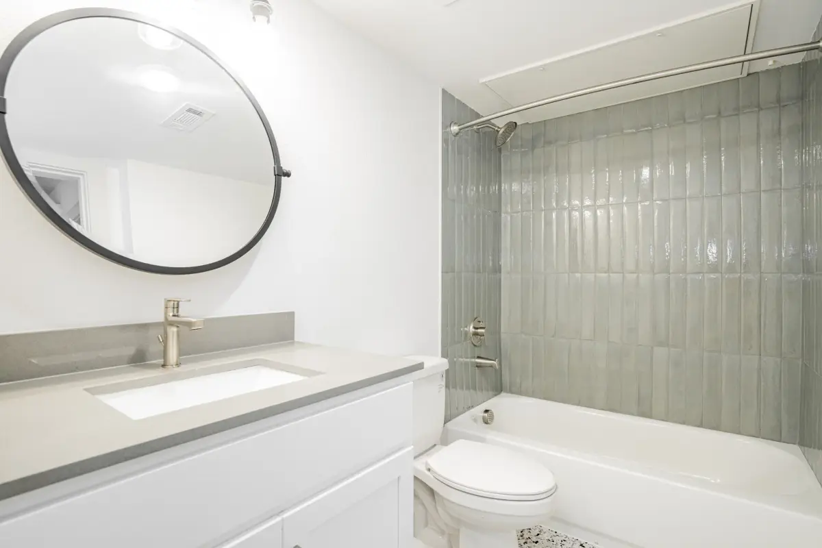 Bathroom with a white vanity, quartz top, round black-framed mirror, and a sage-green tiled tub-and-shower