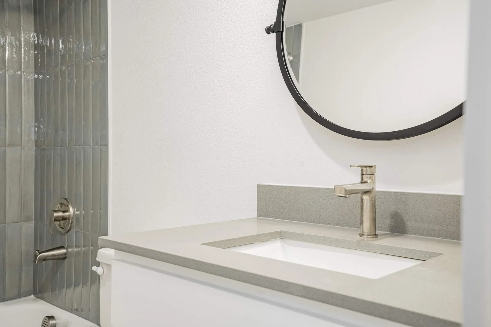 Sage-green tiled tub-and-shower beside a white vanity with a quartz top, brushed-gold faucet, and round black-framed mirror